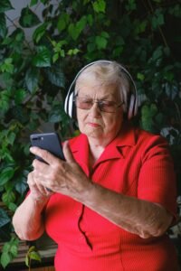 Focused aged female wearing red outfit in eyeglasses and headphones listening to music while browsing cellphone in room with green plants
